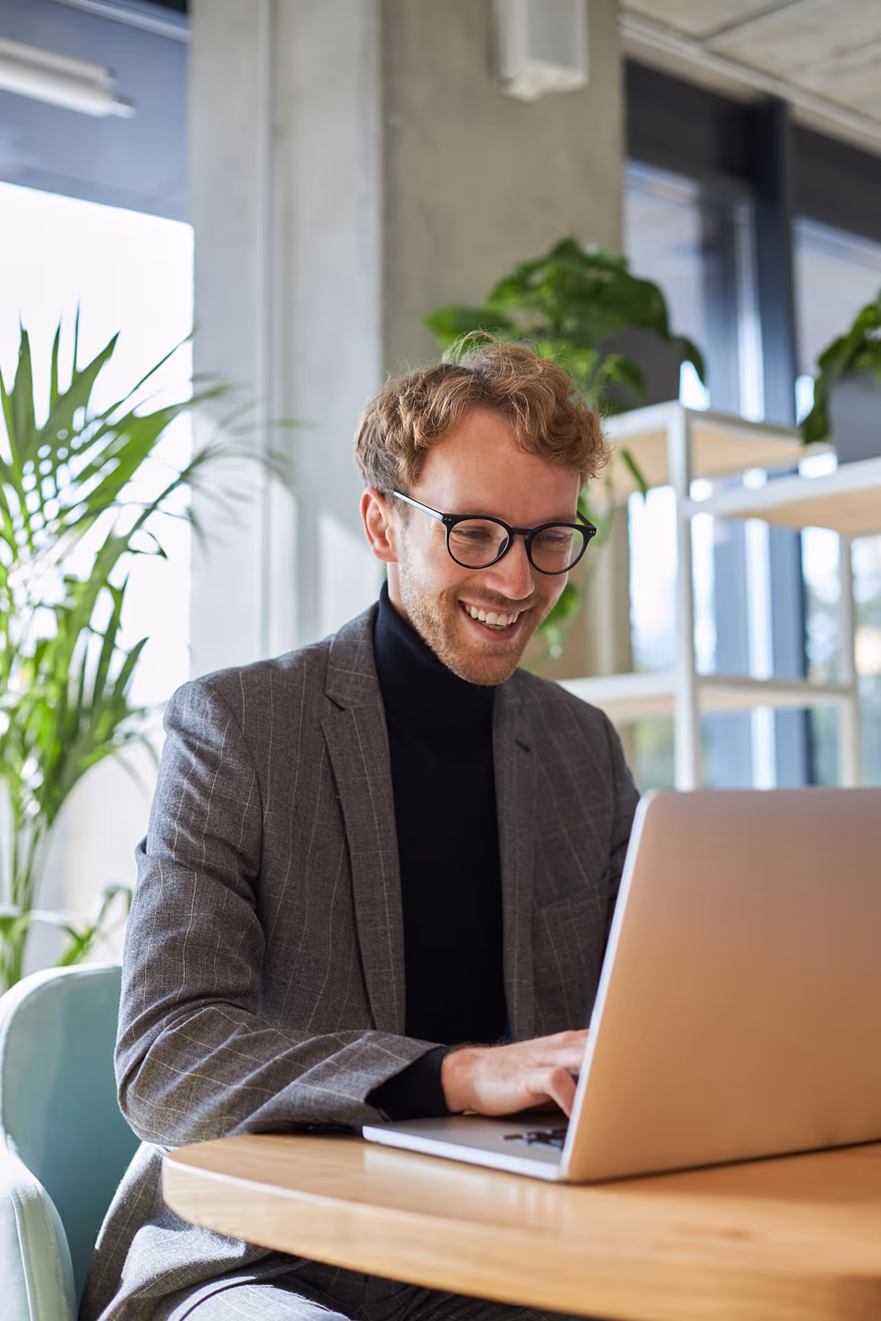 Smiling man with glasses in a gray suit working on a laptop at a wooden table in a bright office with plants.