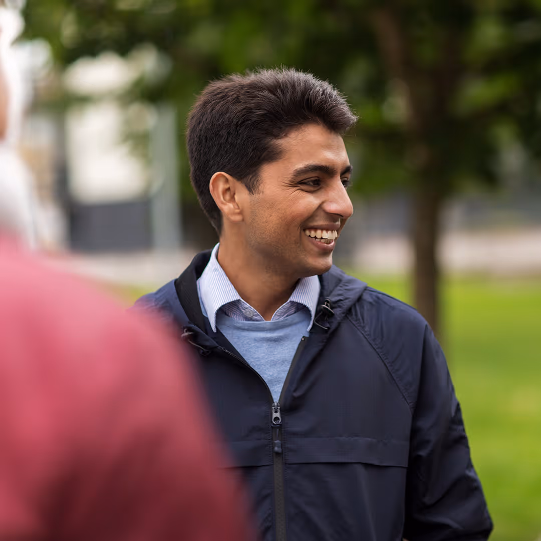 Smiling man in a dark jacket and light blue sweater standing outdoors with blurred greenery in the background.