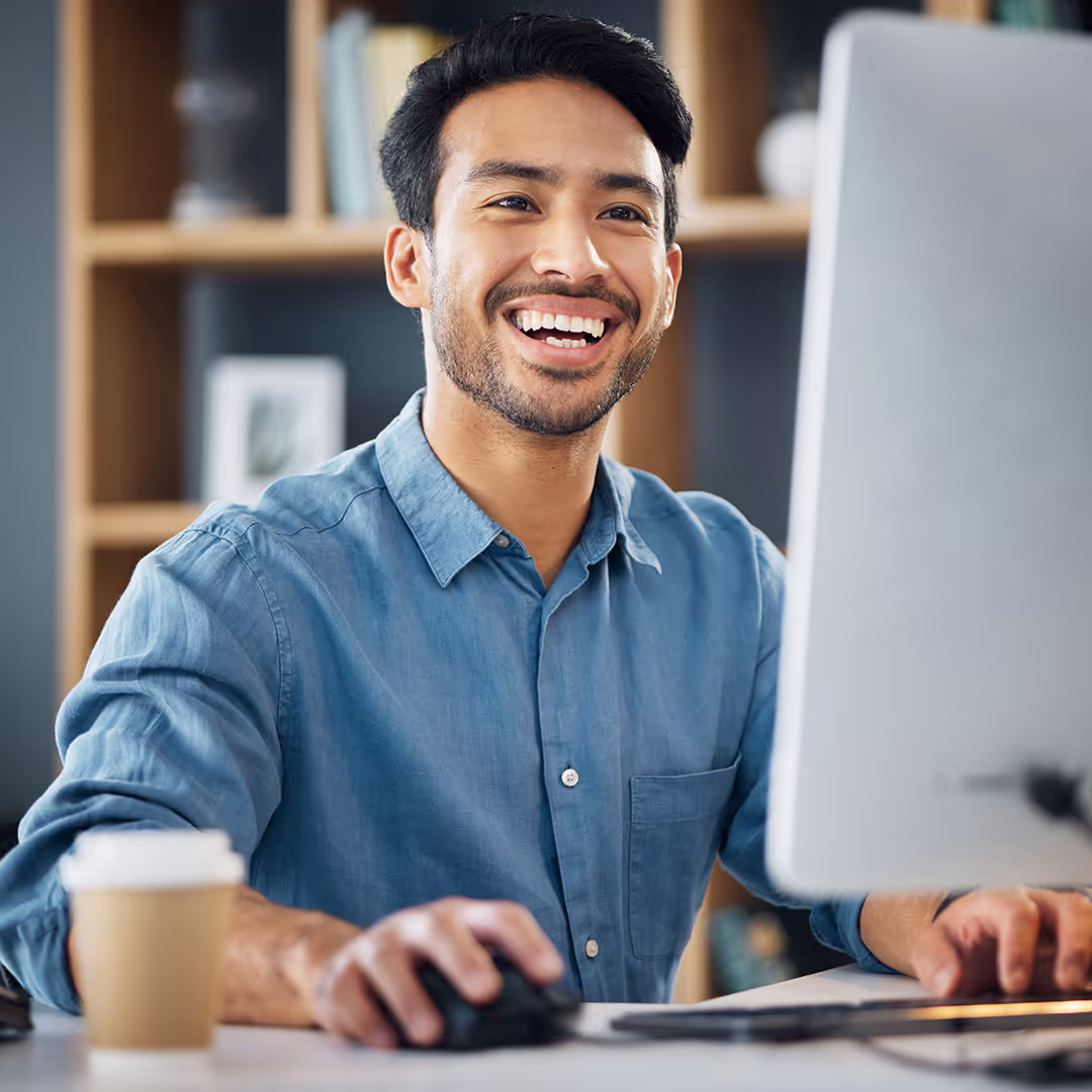 Smiling man in a blue shirt working on a computer in a modern office setting.