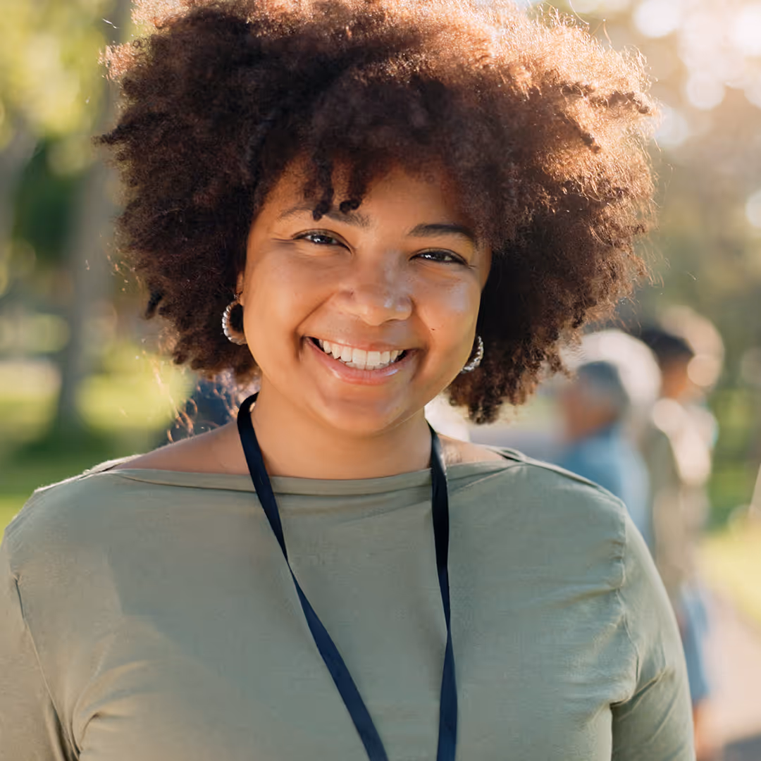 Smiling young woman with curly hair wearing hoop earrings and an olive green top outdoors.
