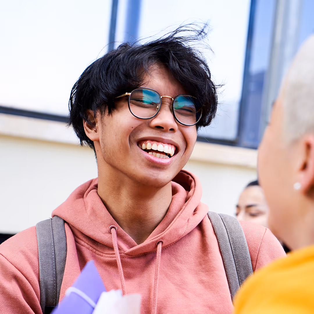 Smiling young person with black hair and glasses wearing a pink hoodie and gray backpack talking to another person.