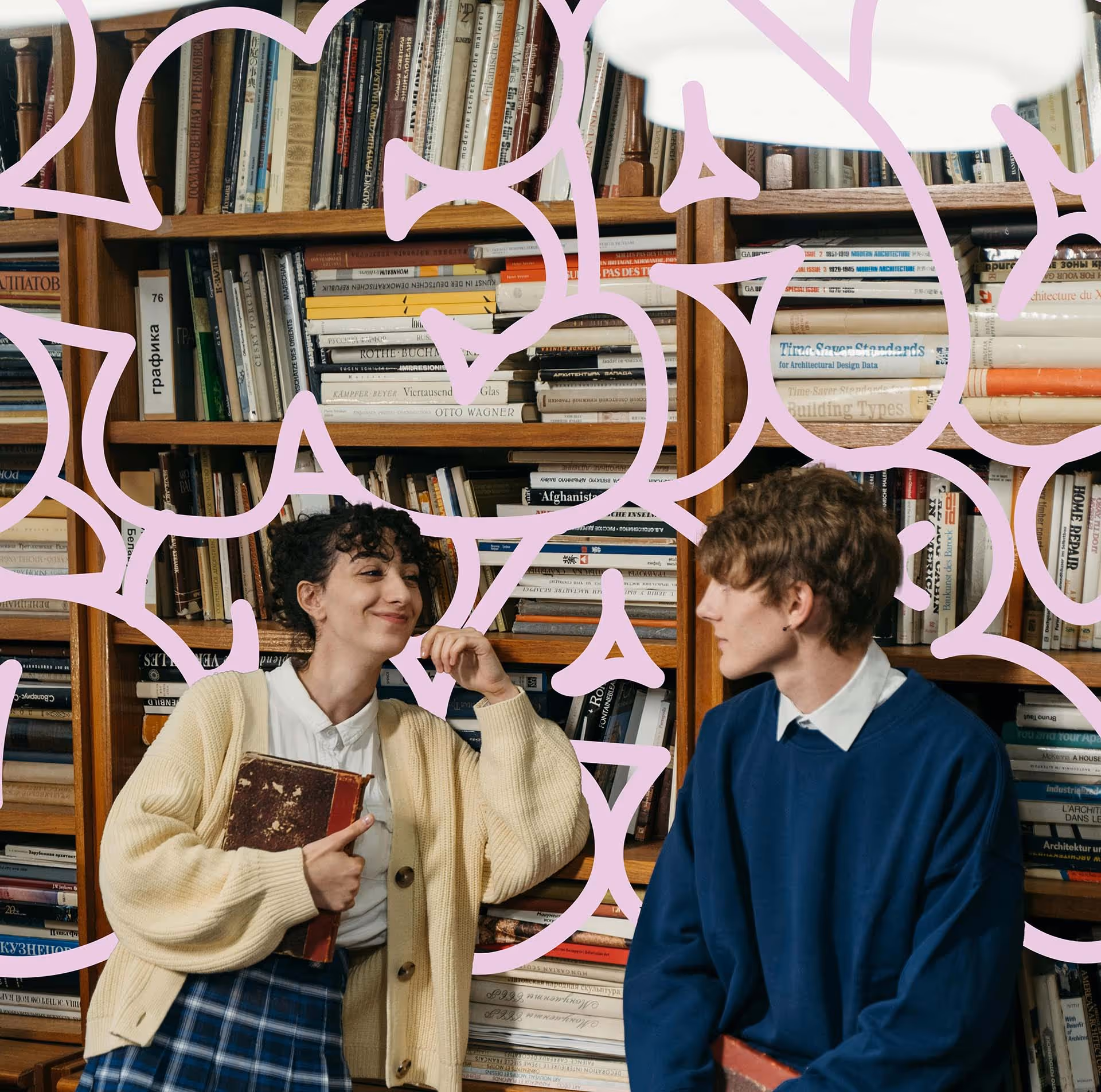 Young woman in a beige cardigan holding a book, smiling and leaning against a bookshelf while a young man in a blue sweater looks at her.