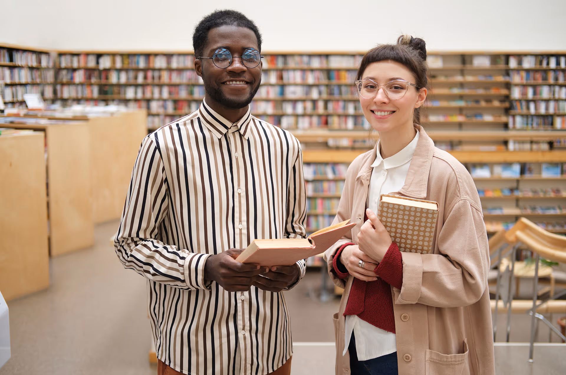 Two young adults smiling in a library, one holding an open book and the other holding a closed book.