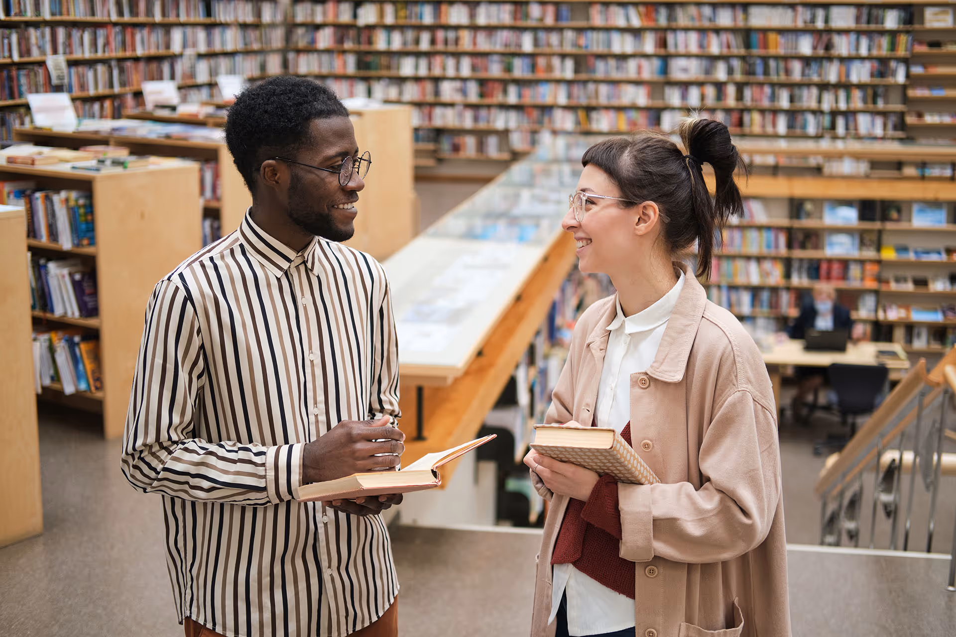 Two students smiling and holding books while talking in a library with shelves full of books in the background.