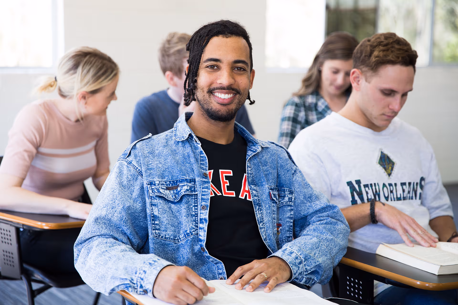 Smiling young man in a denim jacket sitting at a desk with an open book in a classroom with other students.