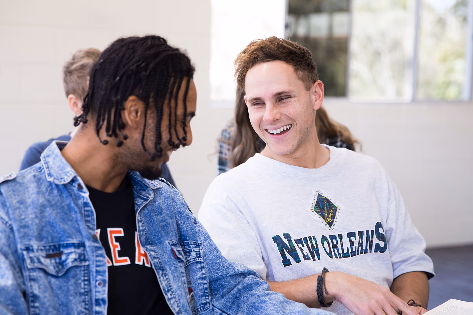 Two young men smiling and talking in a bright indoor setting, one wearing a denim jacket and the other a white New Orleans sweatshirt.