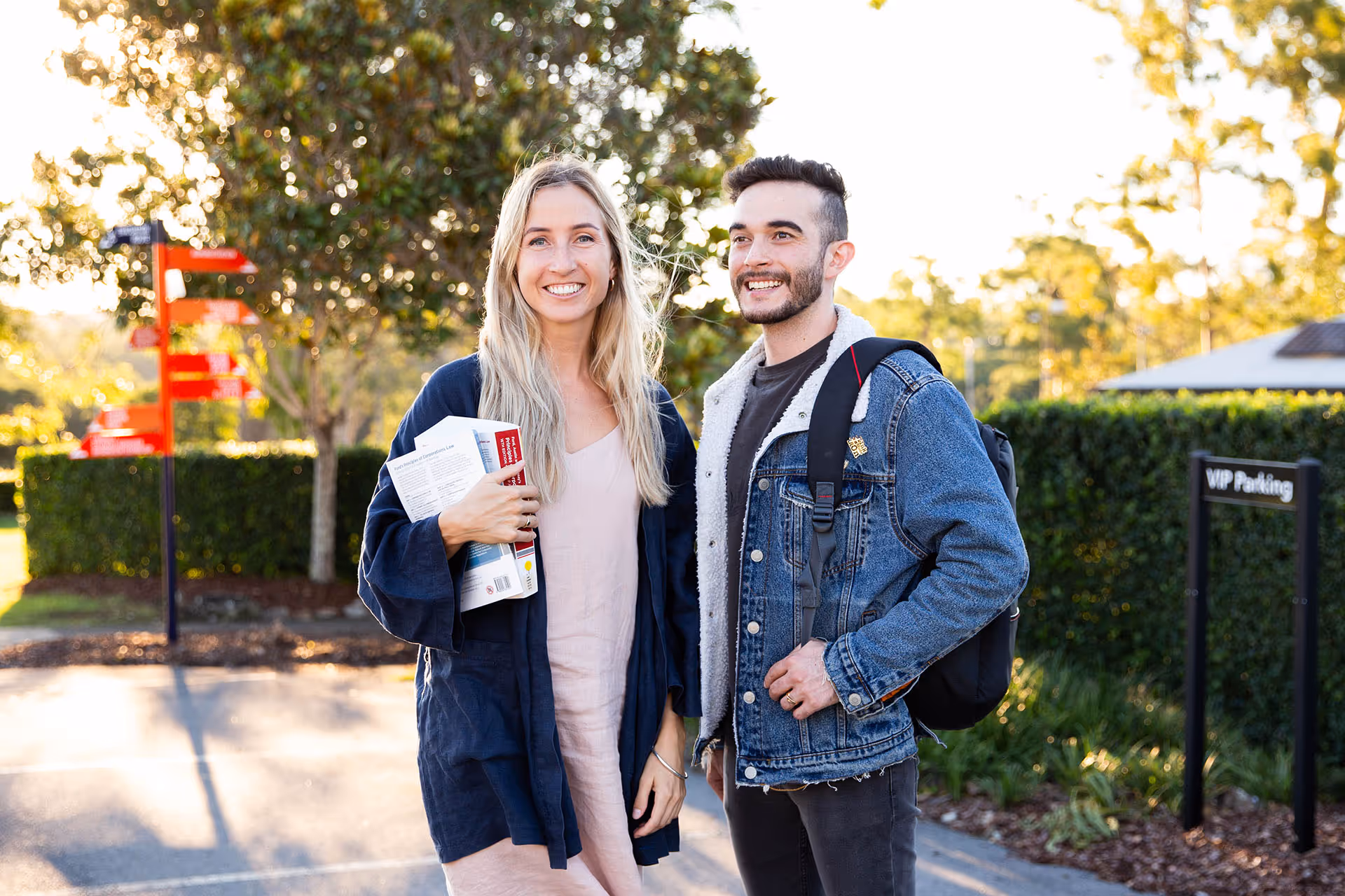 Smiling young woman holding books and a young man with a backpack standing outdoors in a sunlit area.