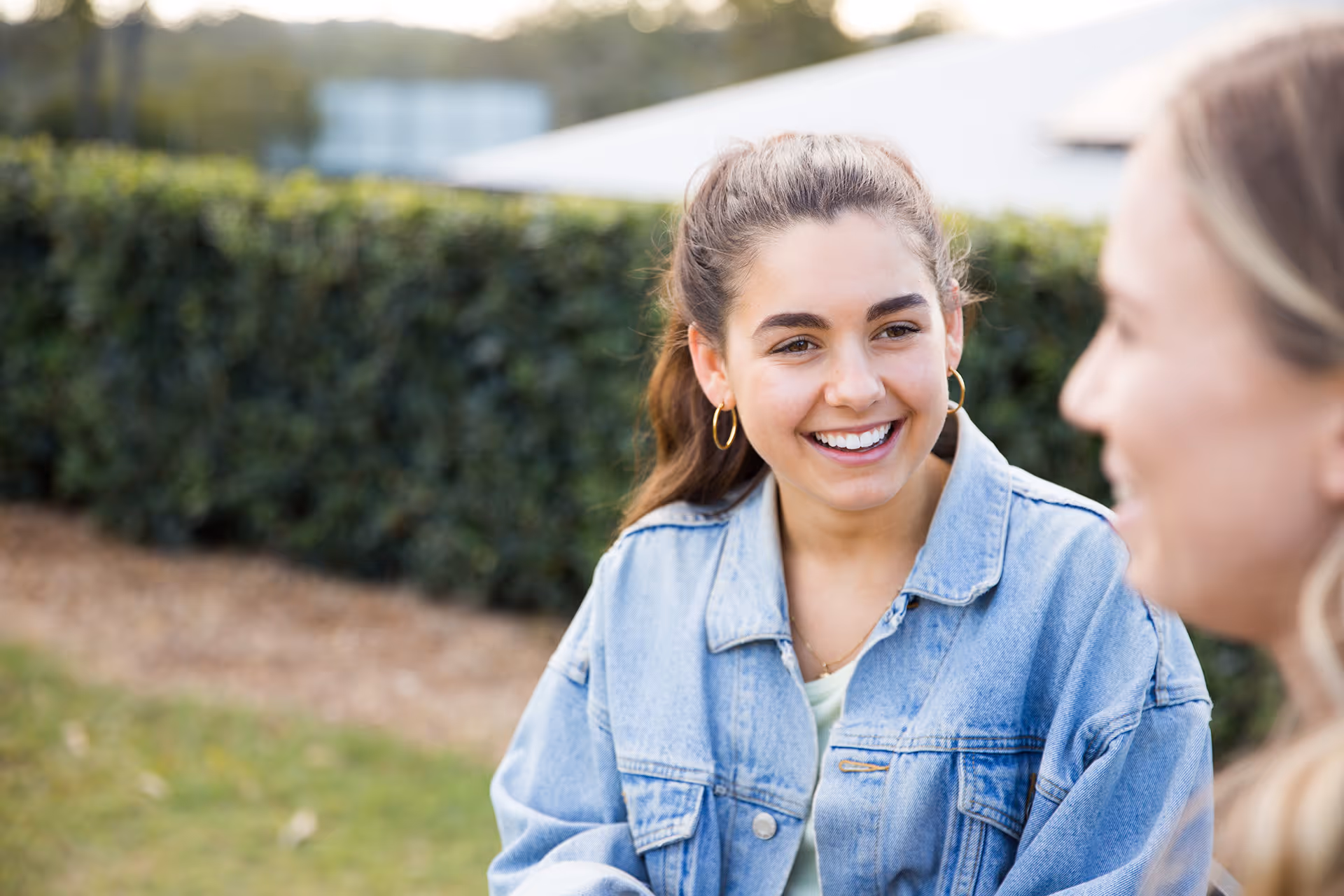 Smiling young woman with ponytail and hoop earrings wearing a denim jacket, talking to another person outdoors.