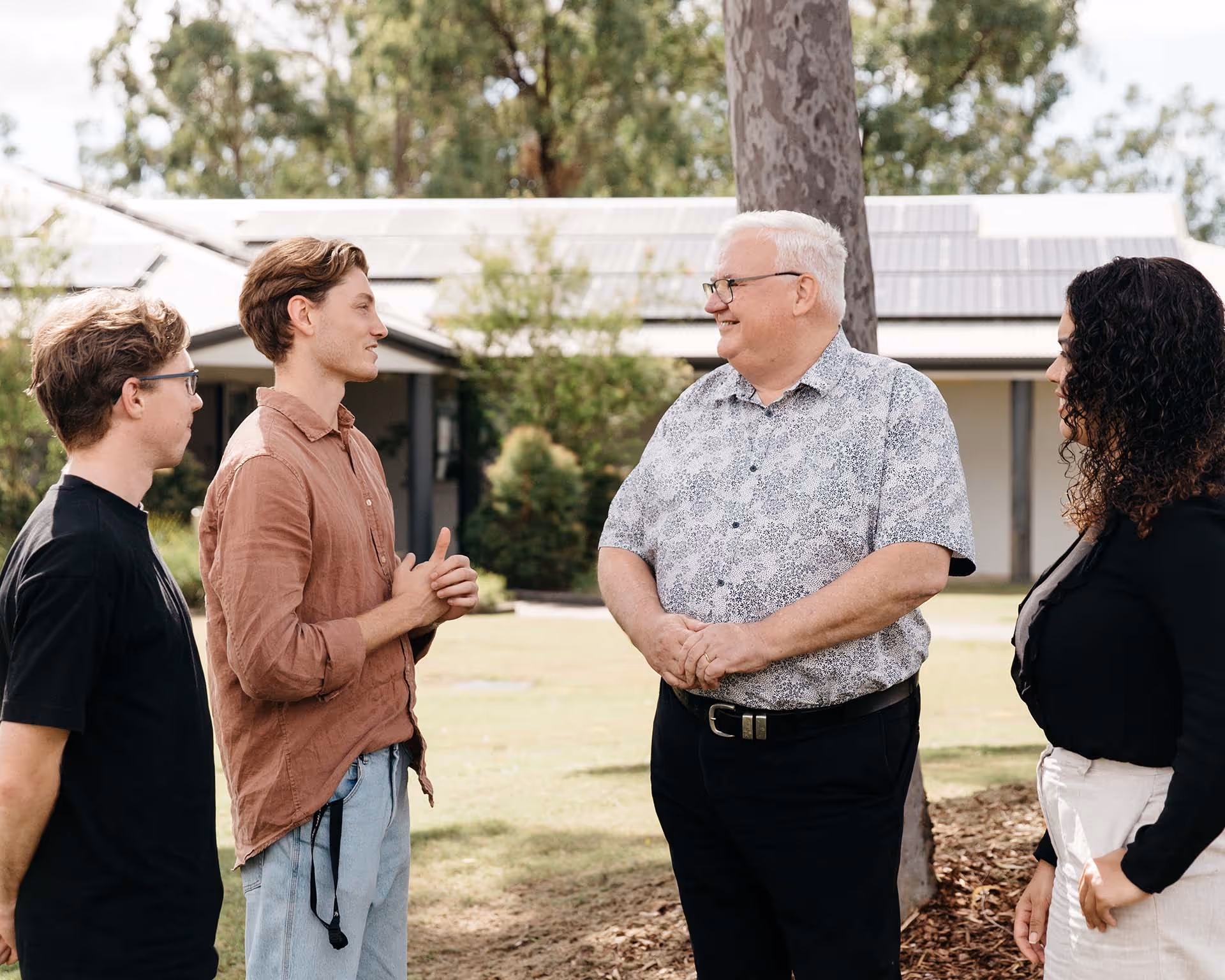Two young men and a woman having a friendly outdoor conversation with an older man near a tree and a building with solar panels.