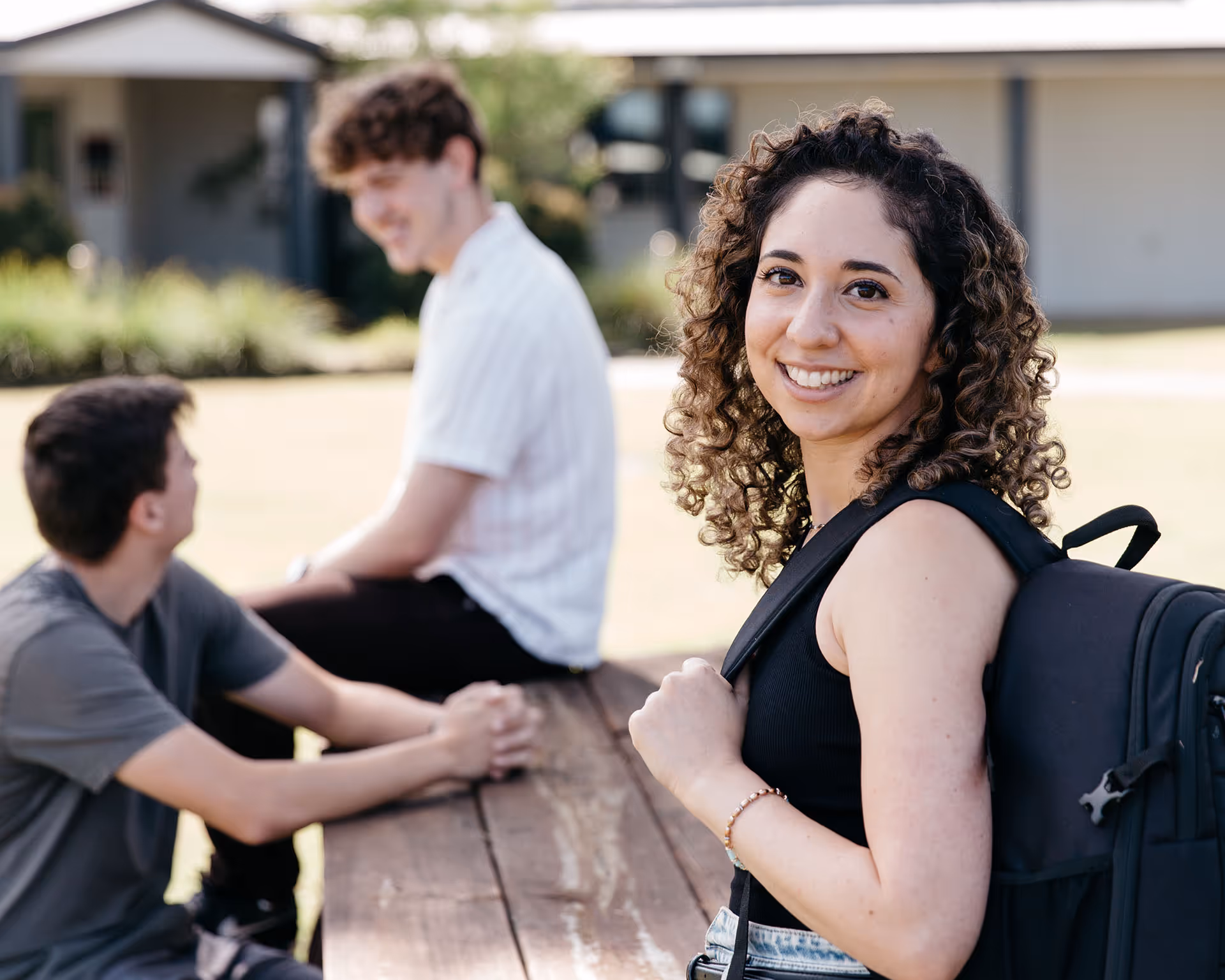 Smiling young woman with curly hair wearing a black backpack standing near a picnic table where two young men are sitting and talking outdoors.