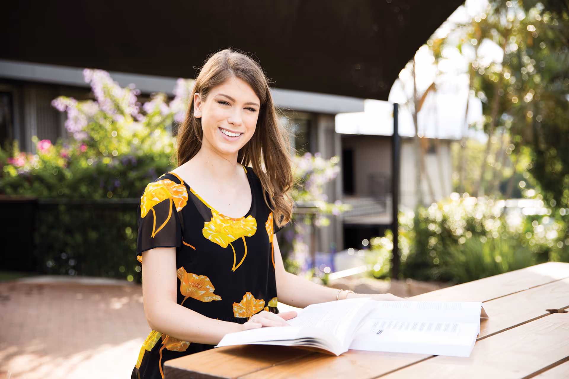 Young woman smiling and sitting at an outdoor wooden table reading a book, with greenery and flowers in the background.