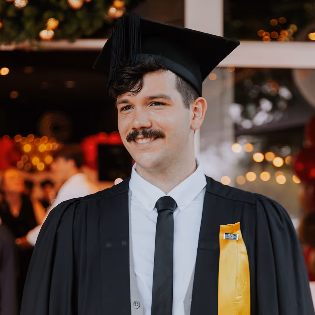 Smiling young man wearing a black graduation cap and gown with a yellow sash at an indoor ceremony.