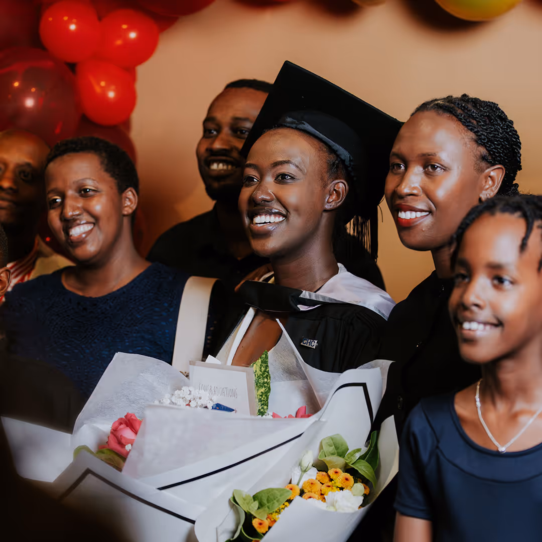 Smiling graduate in a cap and gown holding bouquets, surrounded by happy family and friends.