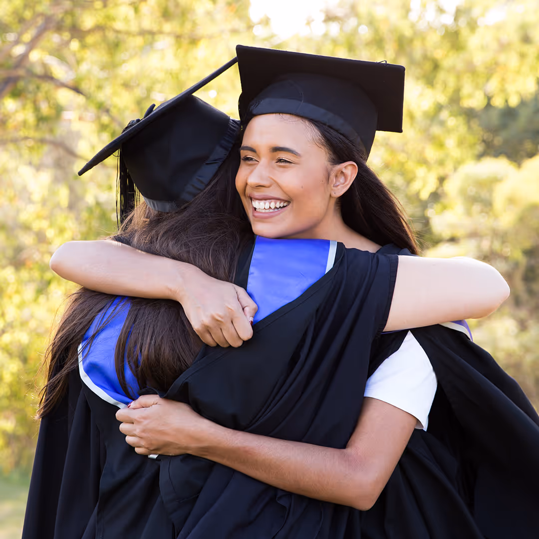 Two graduates wearing caps and gowns hugging outdoors with greenery in the background.