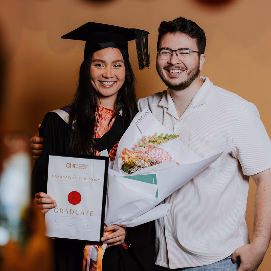 Smiling female graduate in cap and gown holding a diploma next to a man holding a bouquet of flowers.