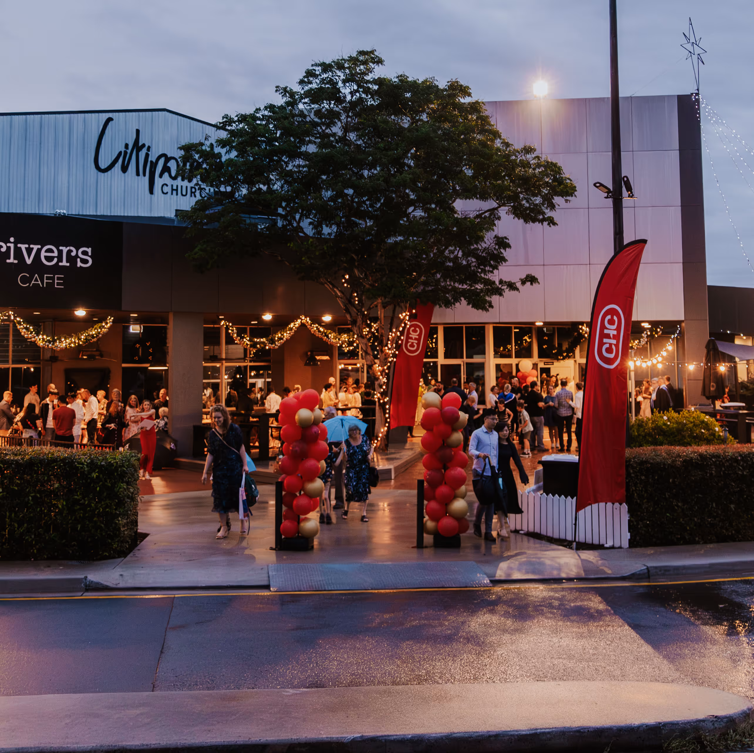 Evening crowd entering a lit venue decorated with red and gold balloons and string lights at Citipointe Church.