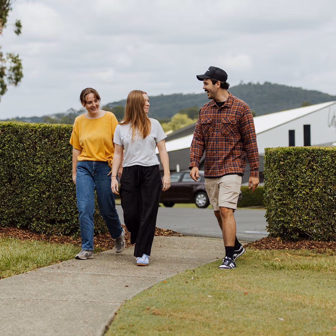 Three young adults walking and talking on a paved path surrounded by green bushes and grass.