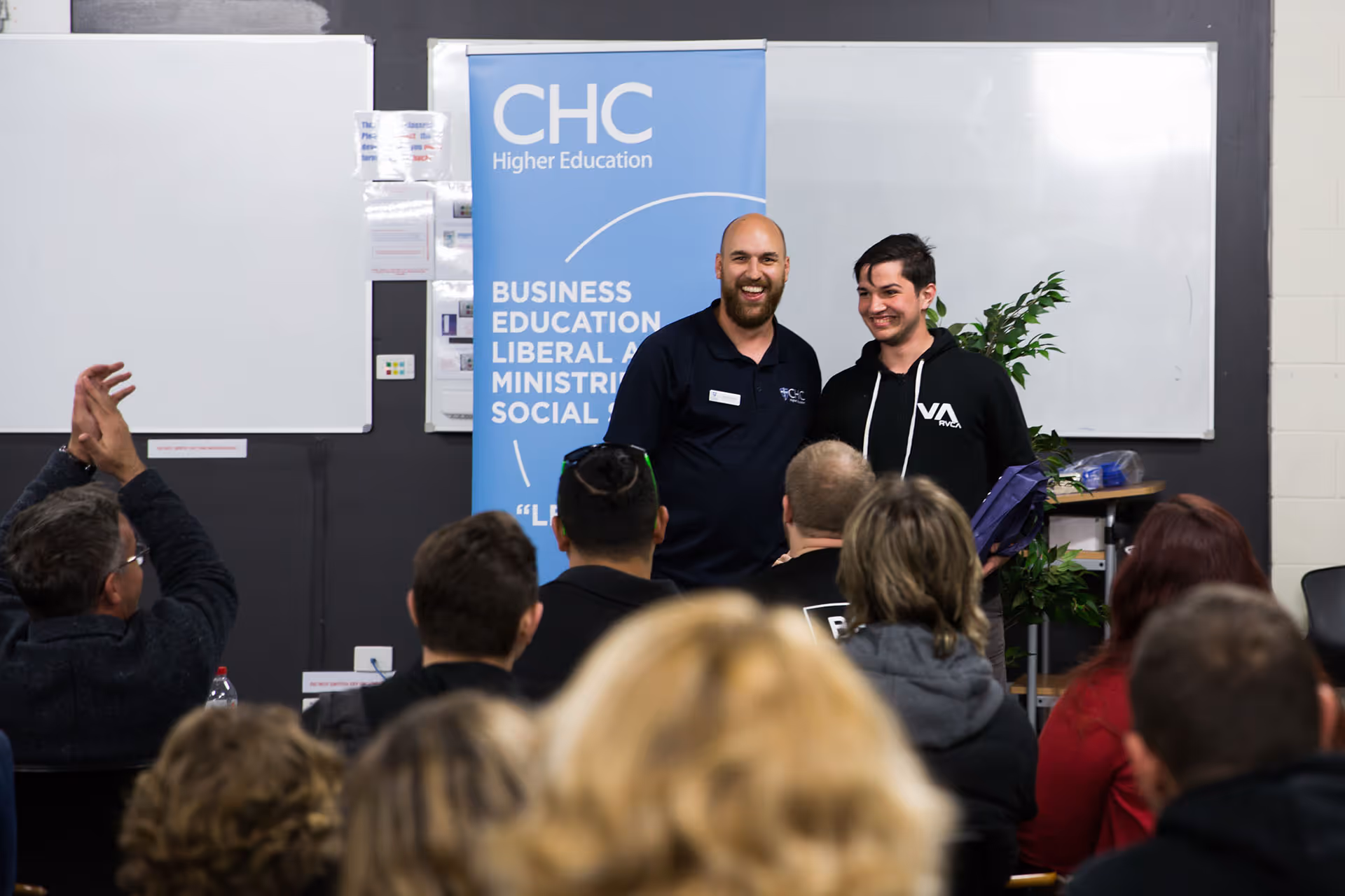 Two men smiling and standing in front of a CHC Higher Education banner while an audience watches and applauds.