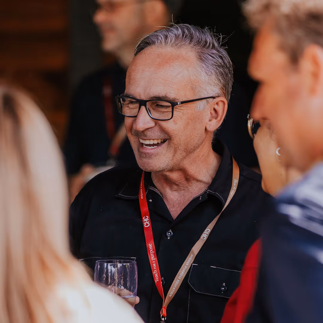 Smiling middle-aged man with gray hair and glasses holding a glass and wearing lanyards at a social event.