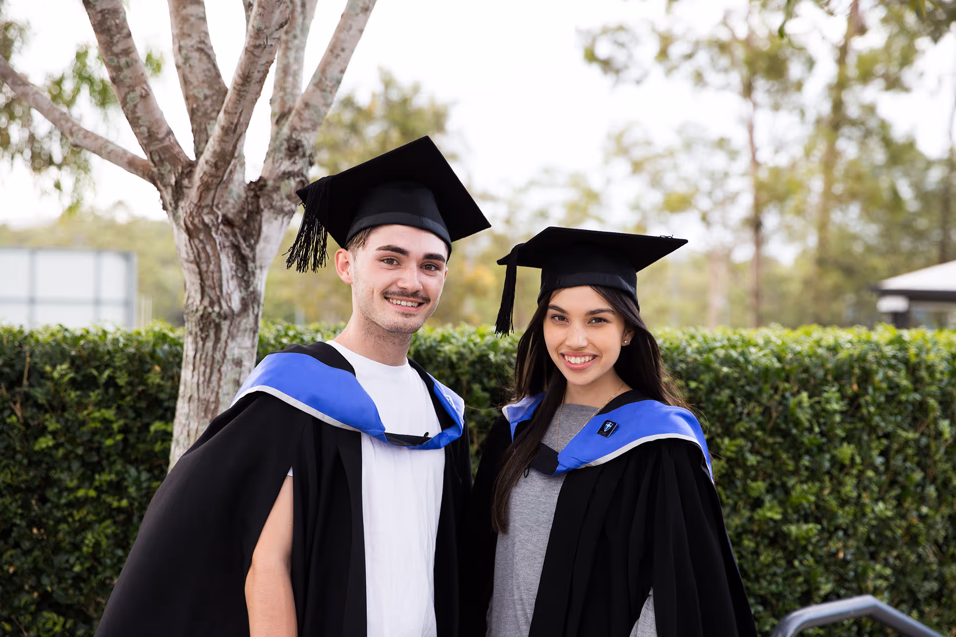 Smiling young man and woman wearing graduation caps and gowns standing outdoors with greenery in the background.