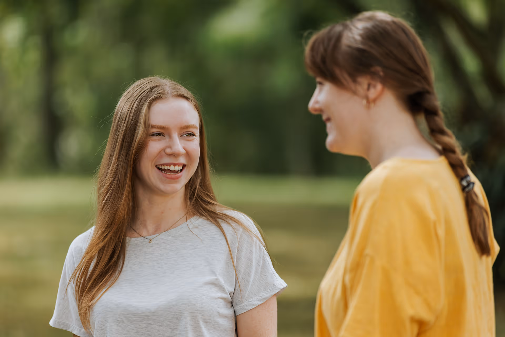 Two women outdoors, one smiling and laughing wearing a white shirt, the other in a yellow shirt with a braid.