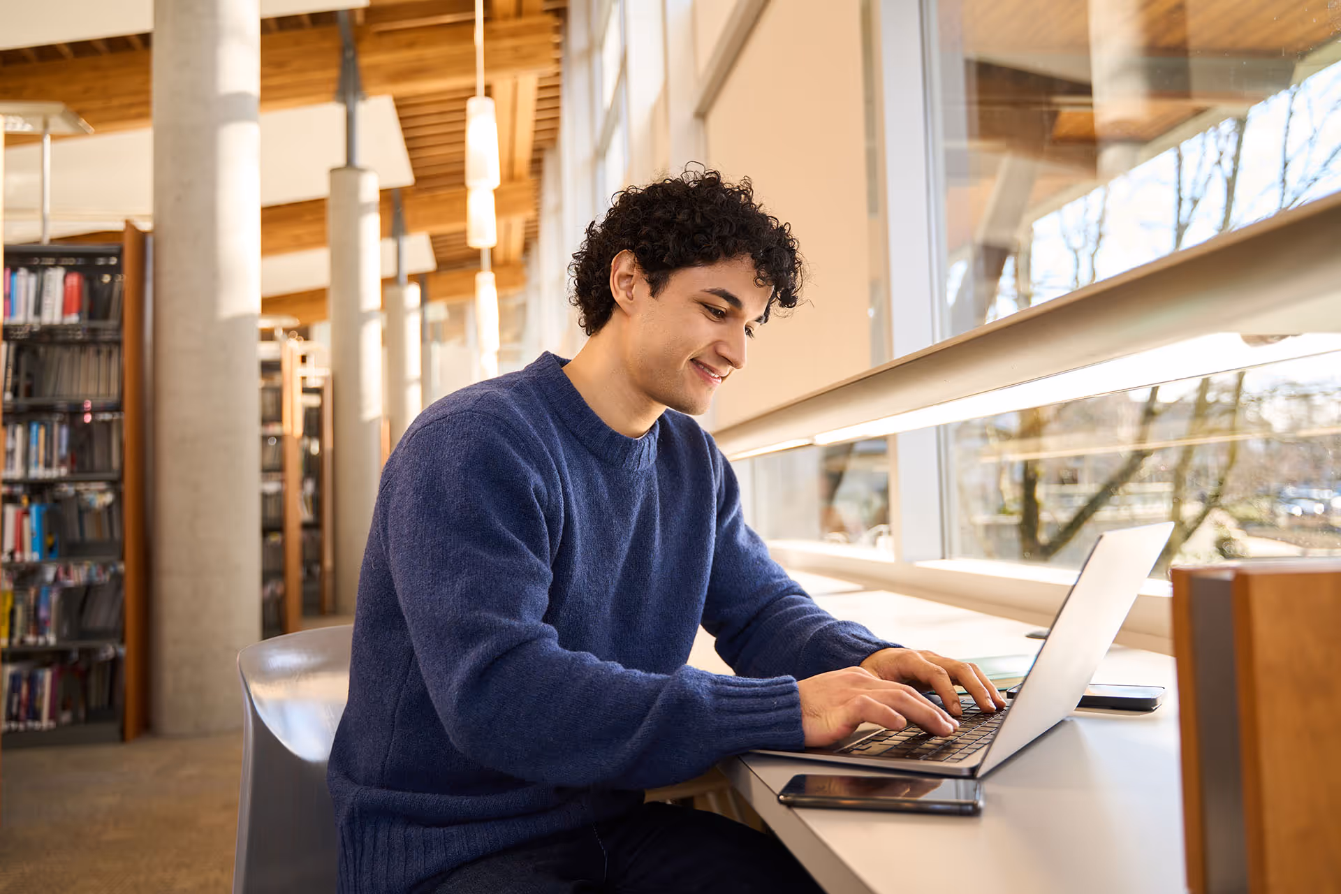 Young man in a blue sweater typing on a laptop at a library desk beside a window.