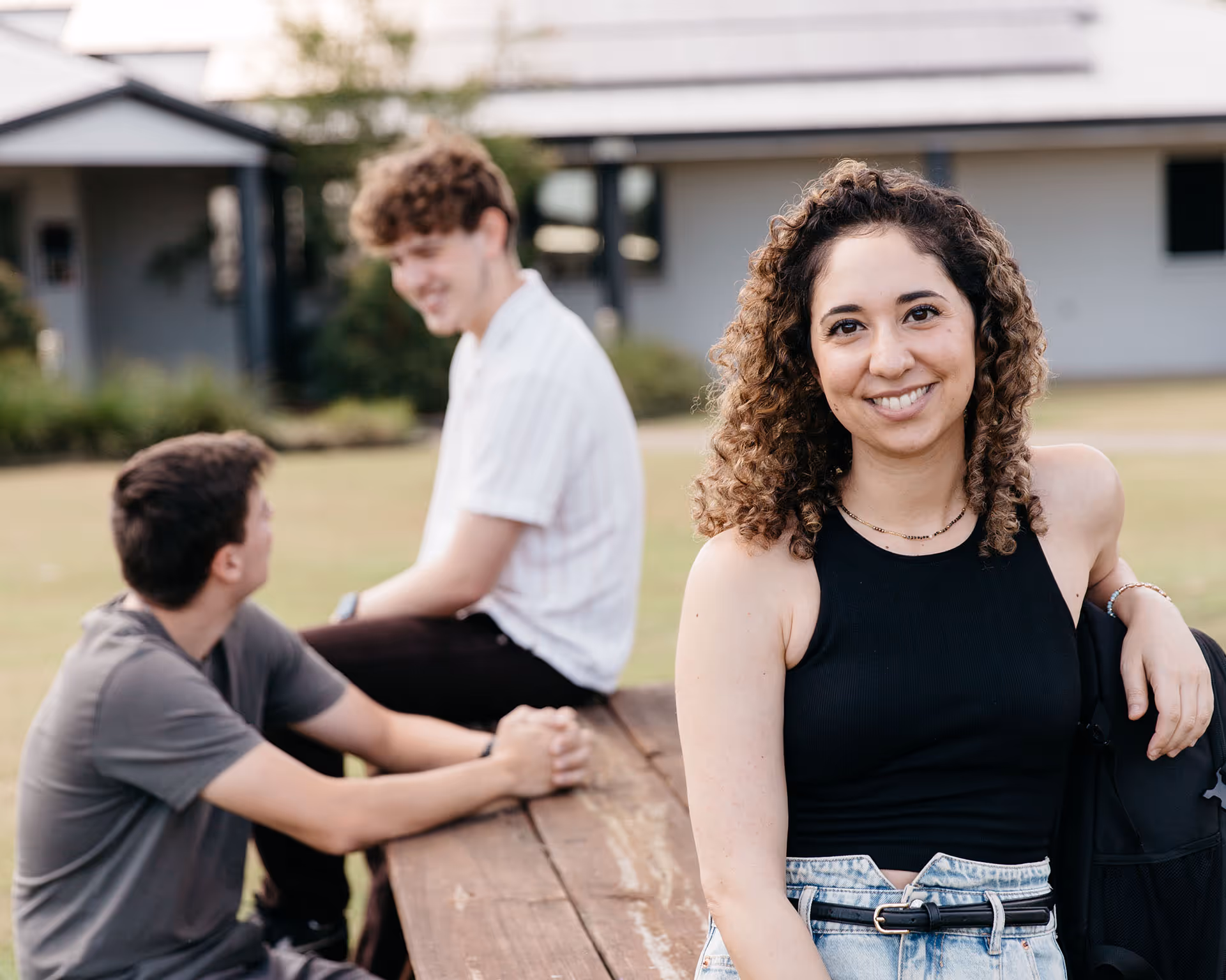 Smiling young woman in black sleeveless top sitting at a wooden picnic table with two young men talking in the background outdoors.