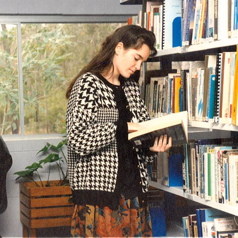 Young woman reading a book beside bookshelves in a library with a window and plant in the background.