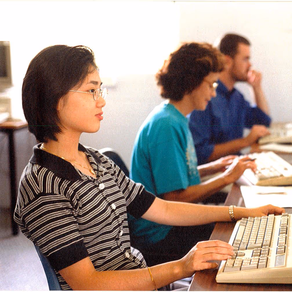 Three young adults sitting in a row at desks using desktop computers with keyboards.