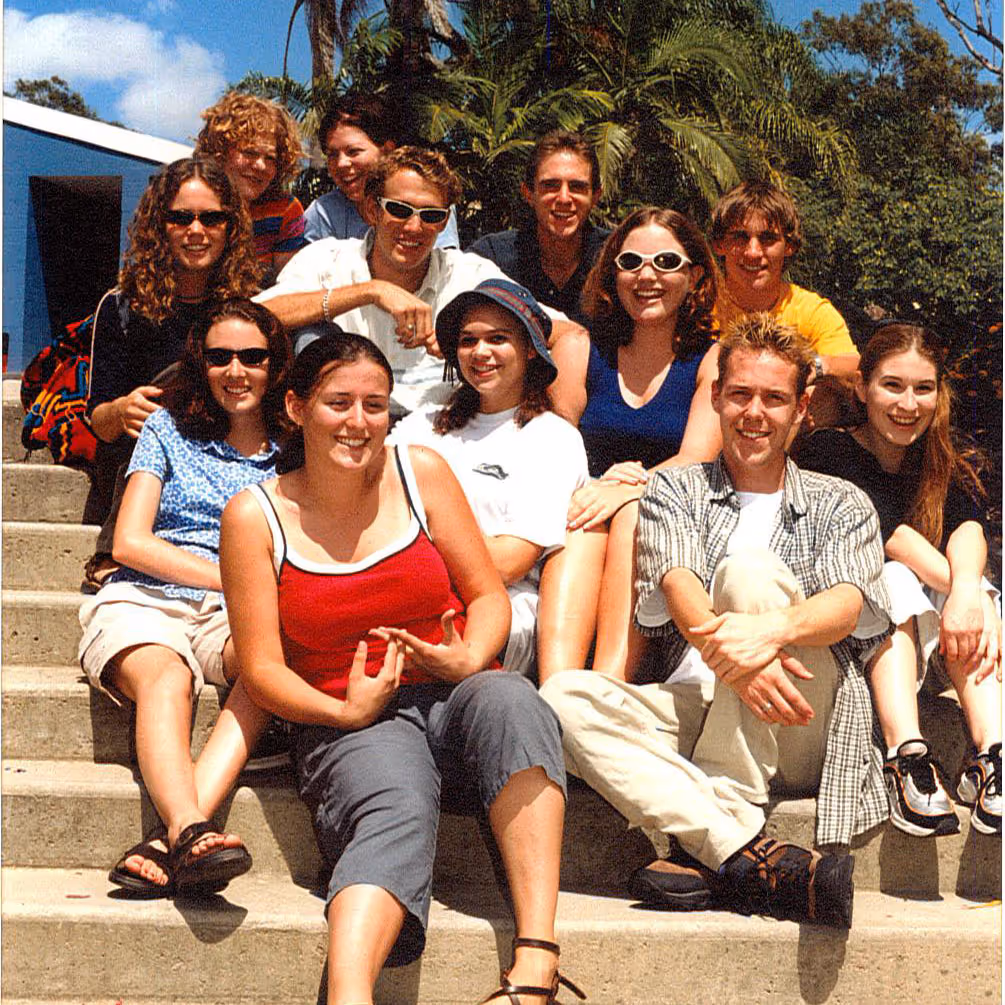 Group of eleven smiling young adults sitting on outdoor concrete steps with trees and blue sky in the background.