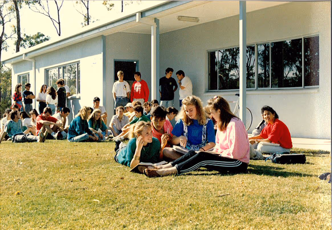 Group of students sitting and standing on grass outside a school building on a sunny day.