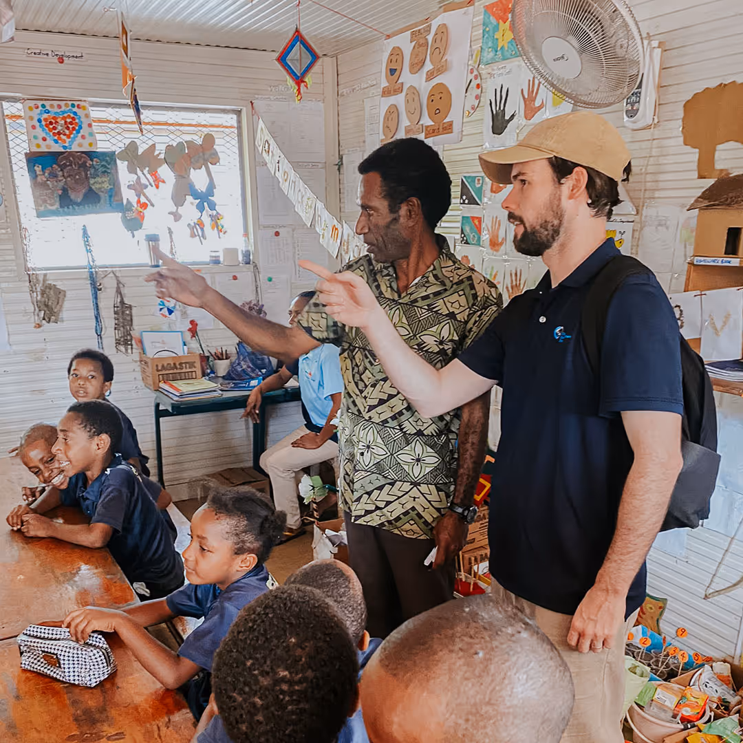 A man and a teacher in a classroom surrounded by children seated at a table, with colorful educational decorations on the walls.