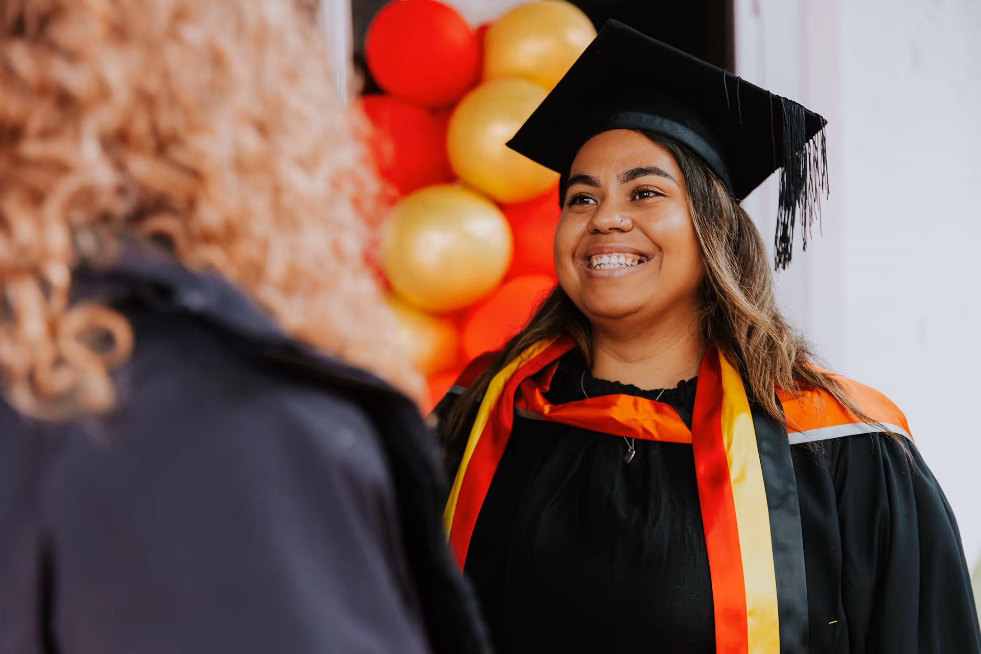 Smiling graduate wearing a black cap and gown with red, yellow, and black sash, talking to another person with curly hair.
