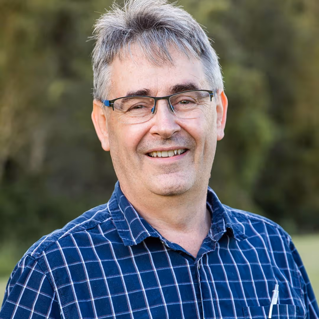 Smiling middle-aged man with gray hair and glasses wearing a blue checkered shirt outdoors.