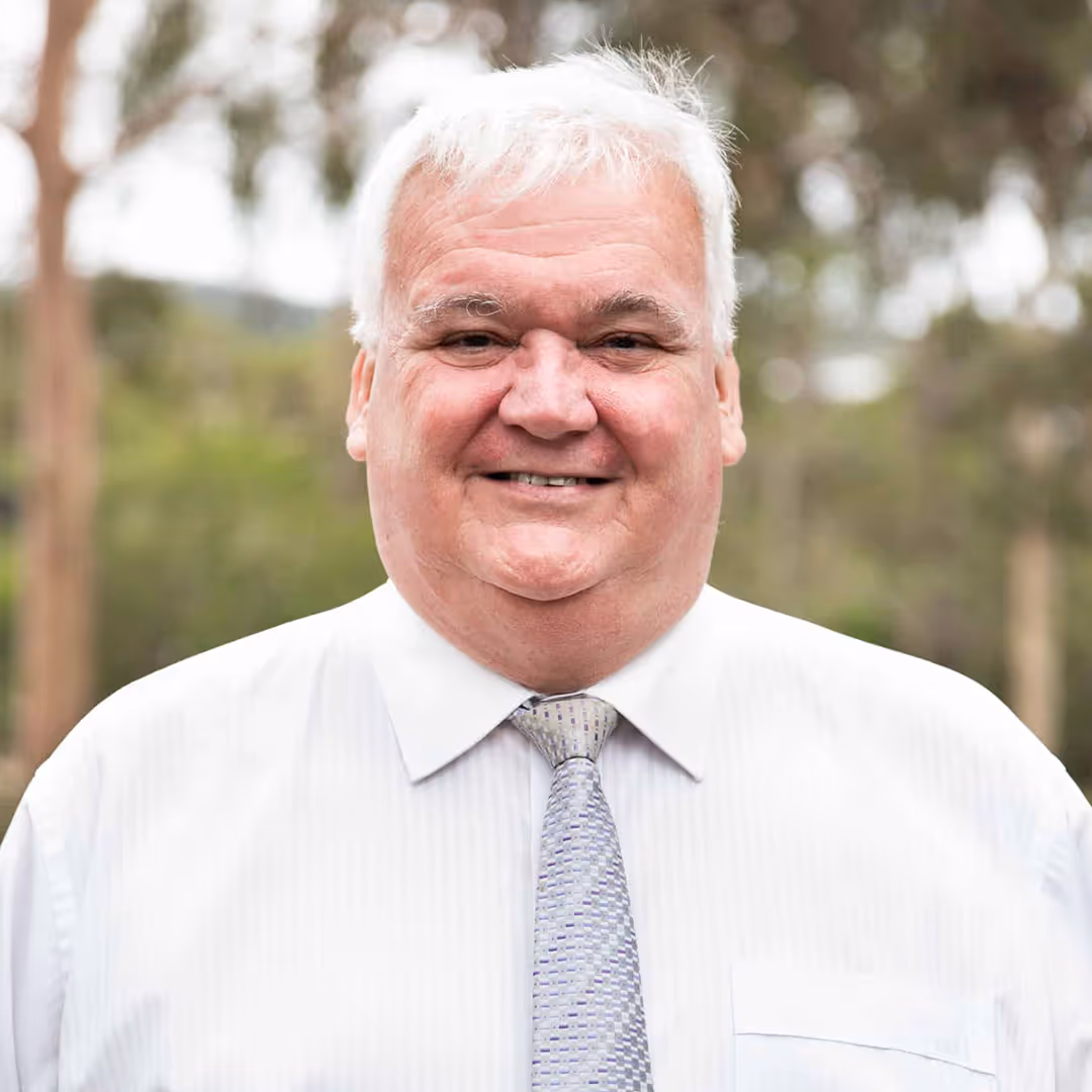 Smiling older man with white hair wearing a white dress shirt and patterned tie outdoors.