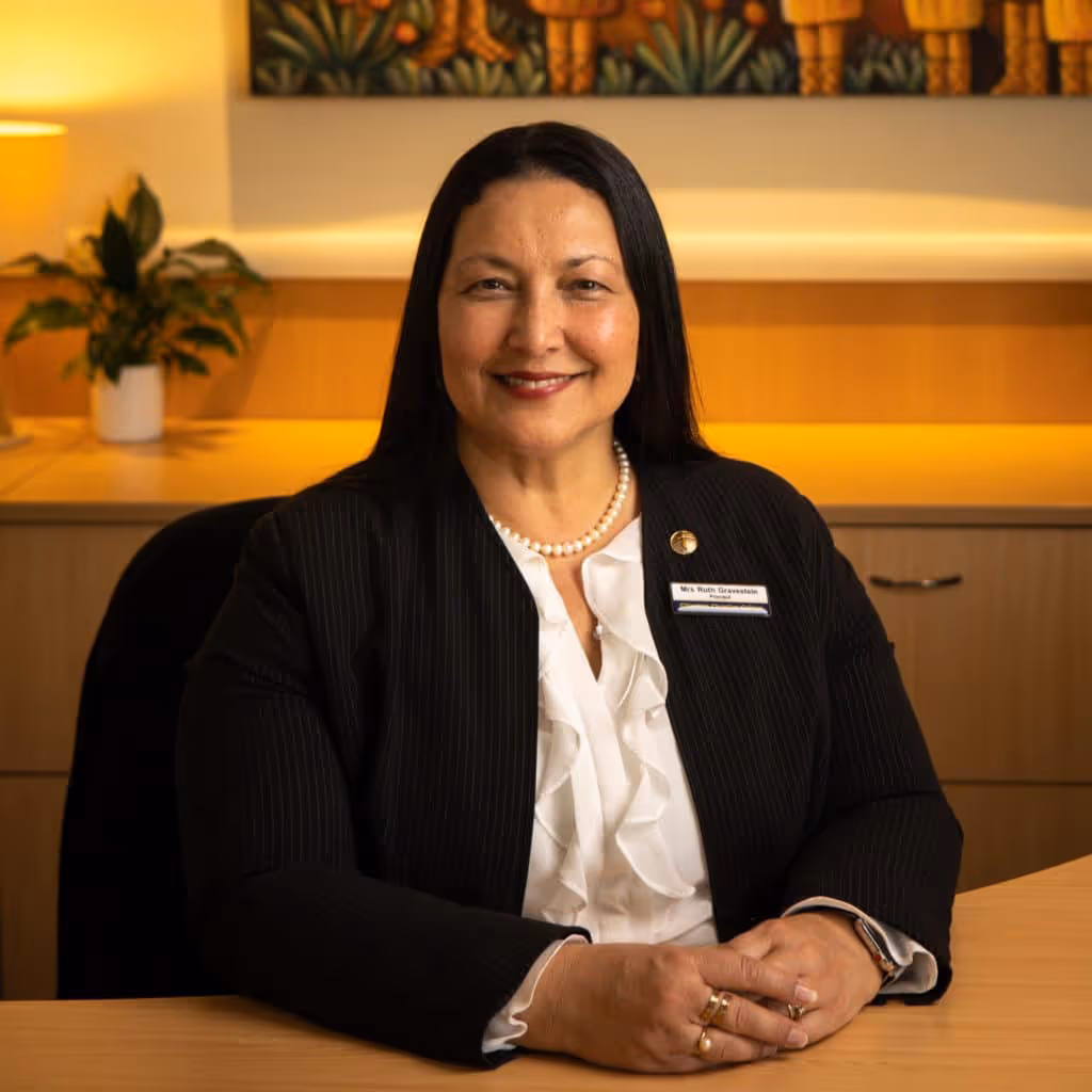 Smiling woman with long dark hair seated at a desk wearing a black blazer and white blouse, with a pearl necklace and a name badge.