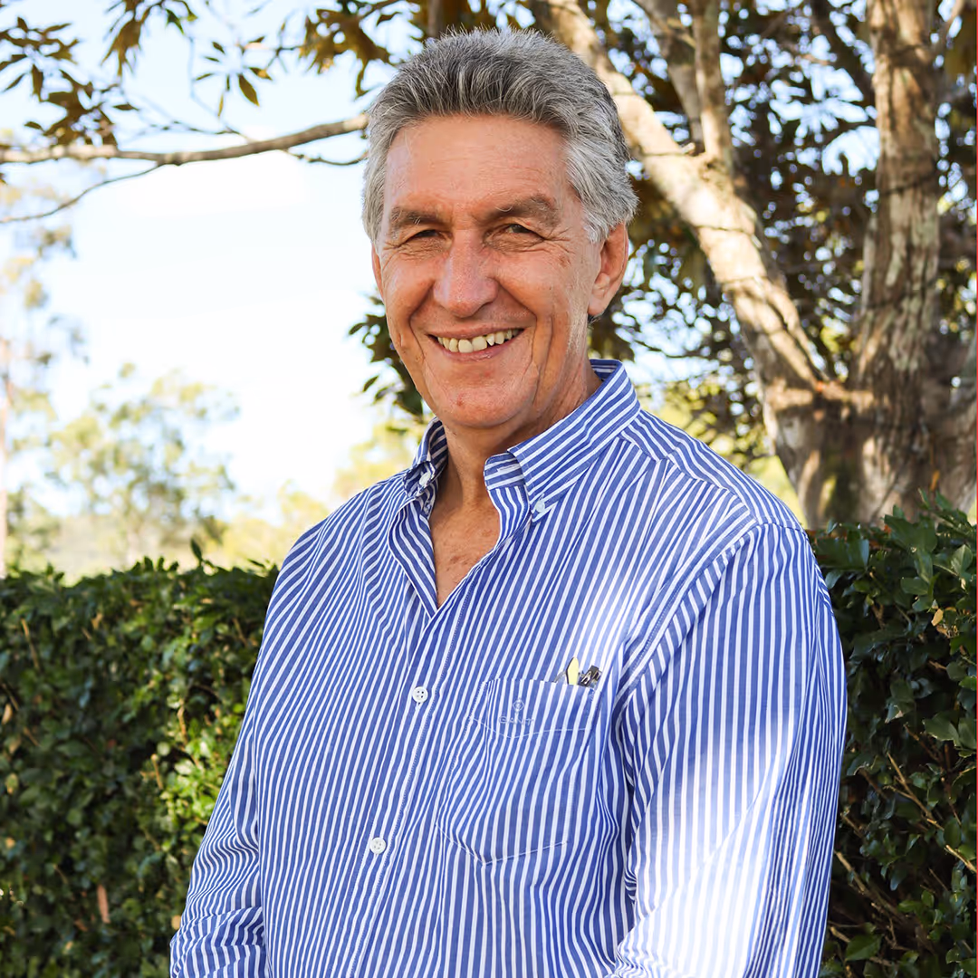 Smiling older man wearing a blue and white striped shirt standing outdoors in front of greenery and trees.