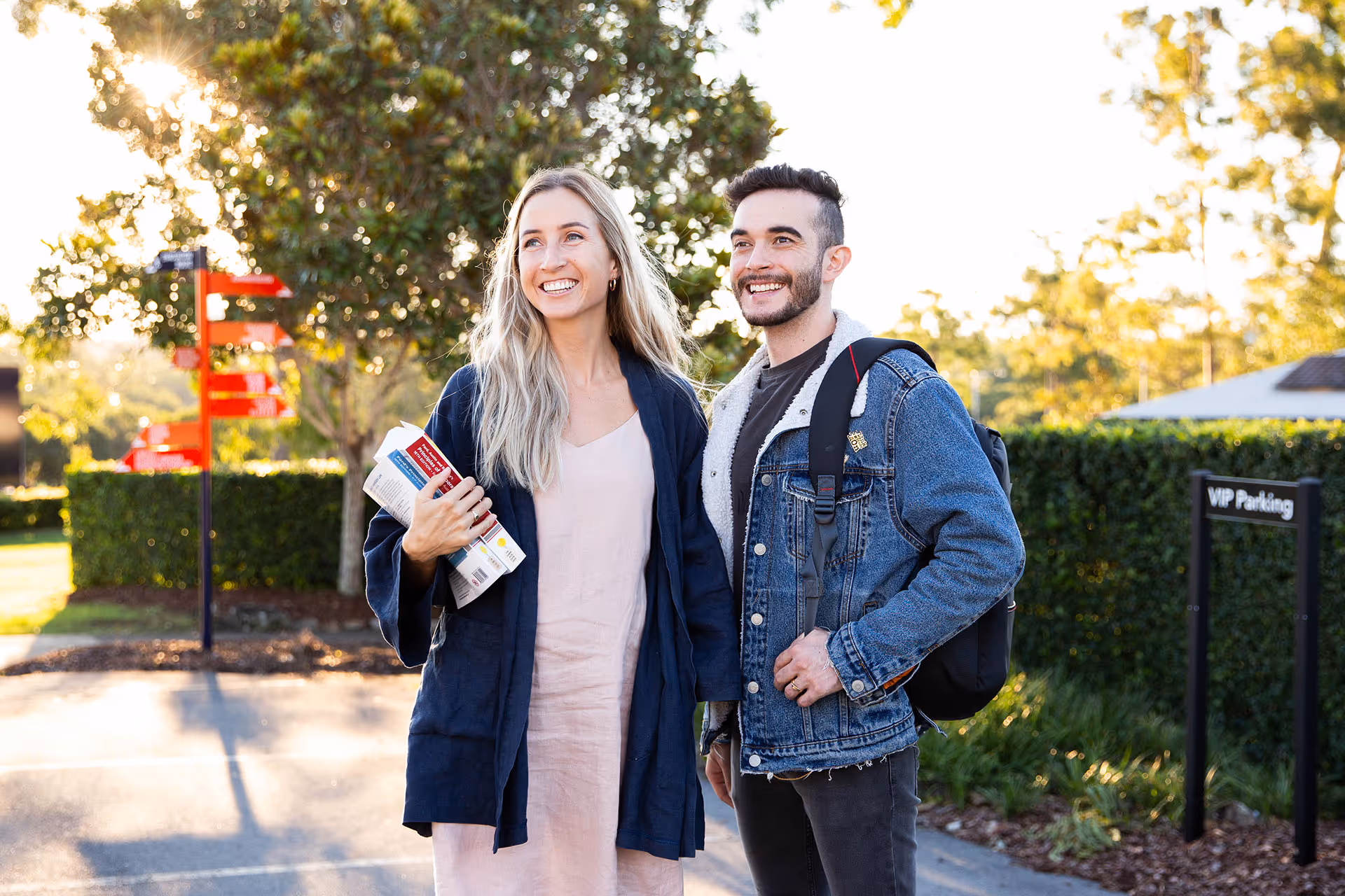 Smiling young woman holding books and man wearing a denim jacket with backpack standing outside near greenery in sunlight.