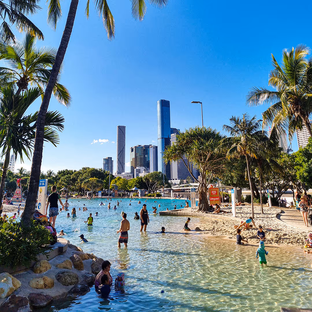 People enjoying a sunny day swimming and playing in a sandy lagoon pool with palm trees and city skyscrapers in the background.