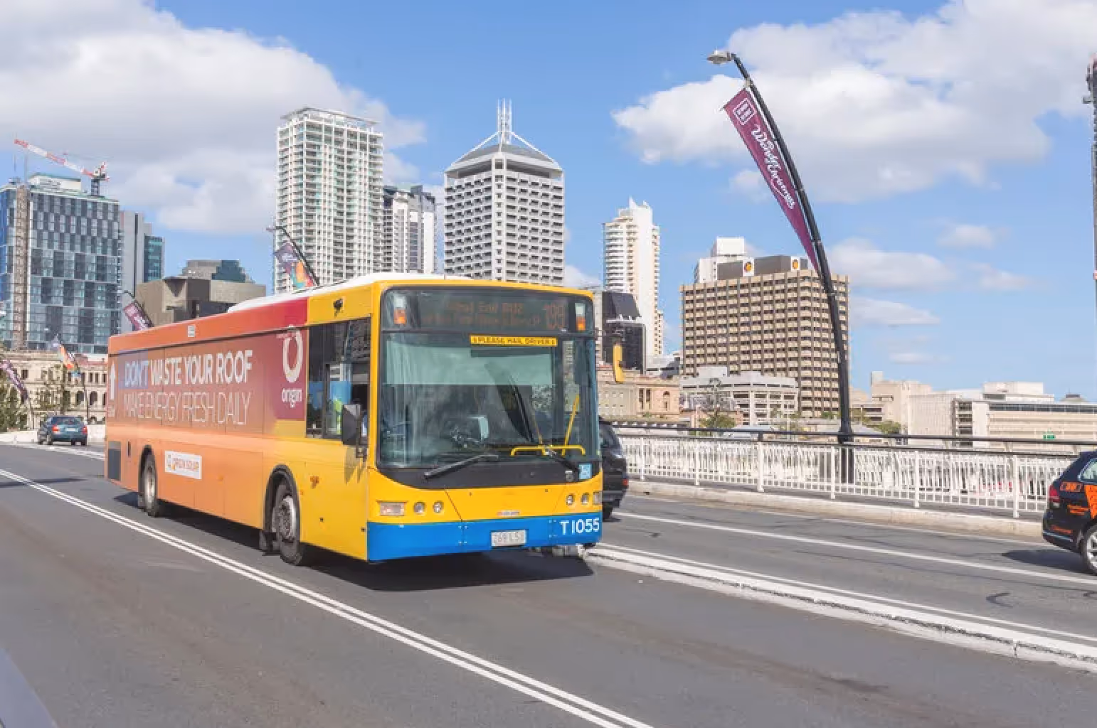 Yellow and orange city bus with advertisement driving on urban road with high-rise buildings in the background.