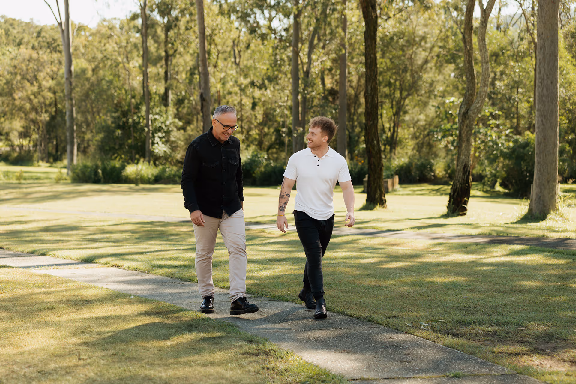 Two men walking on a paved path in a sunlit park surrounded by tall trees and green grass.