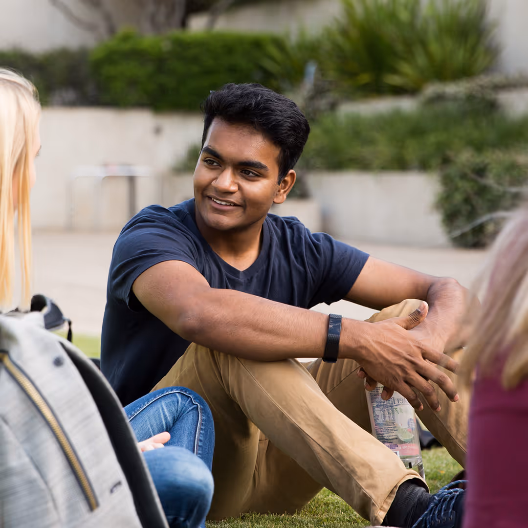 Young man sitting on grass outdoors, smiling and talking with two people partially visible.
