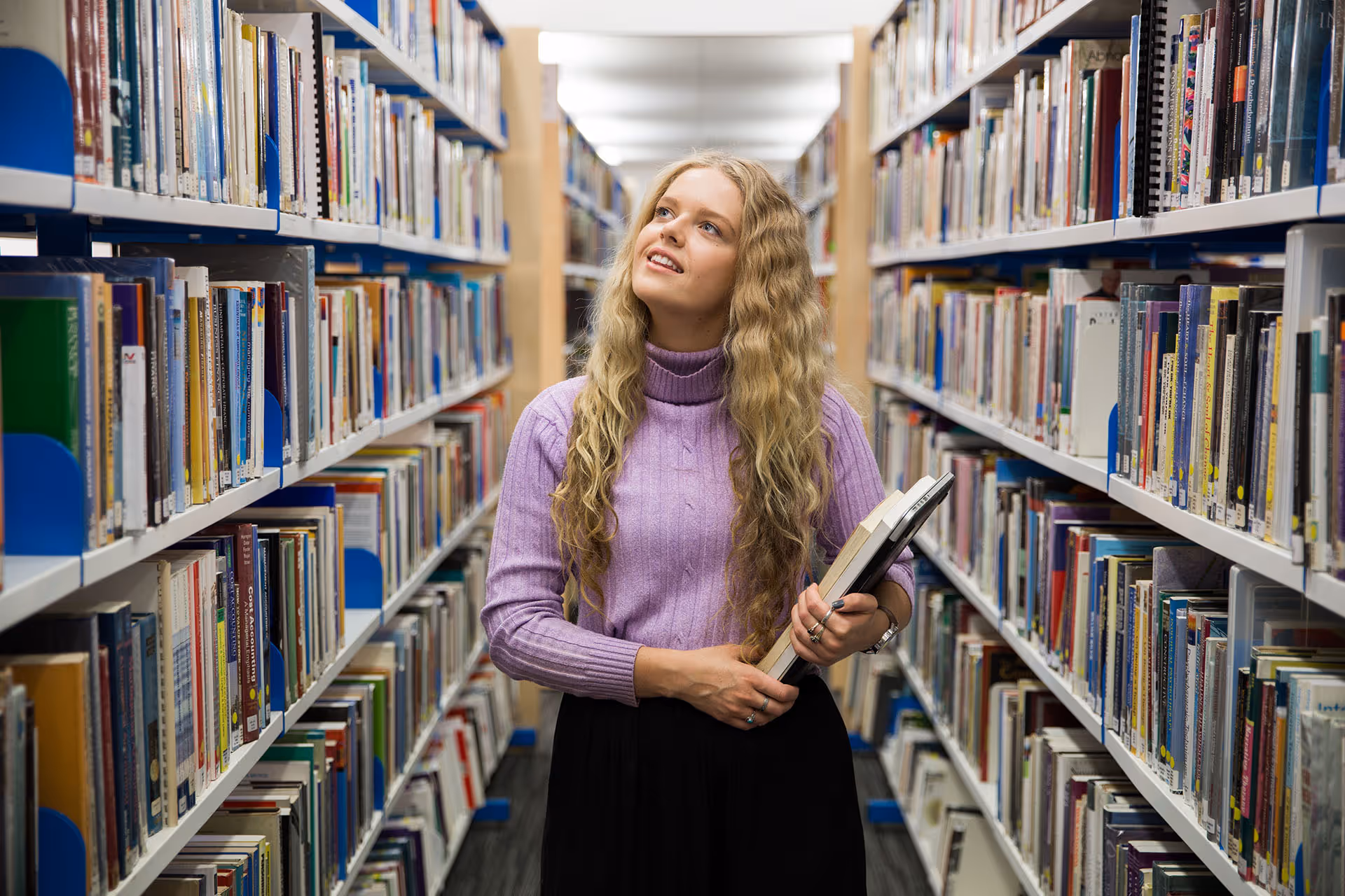 Young woman with long curly hair and a purple sweater holding books in a library aisle.