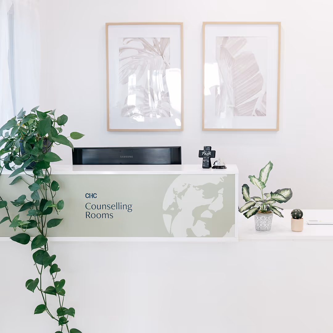 Reception desk labeled 'Counselling Rooms' with a trailing green plant on the left, two framed botanical prints on the wall, and small potted plants on the desk.