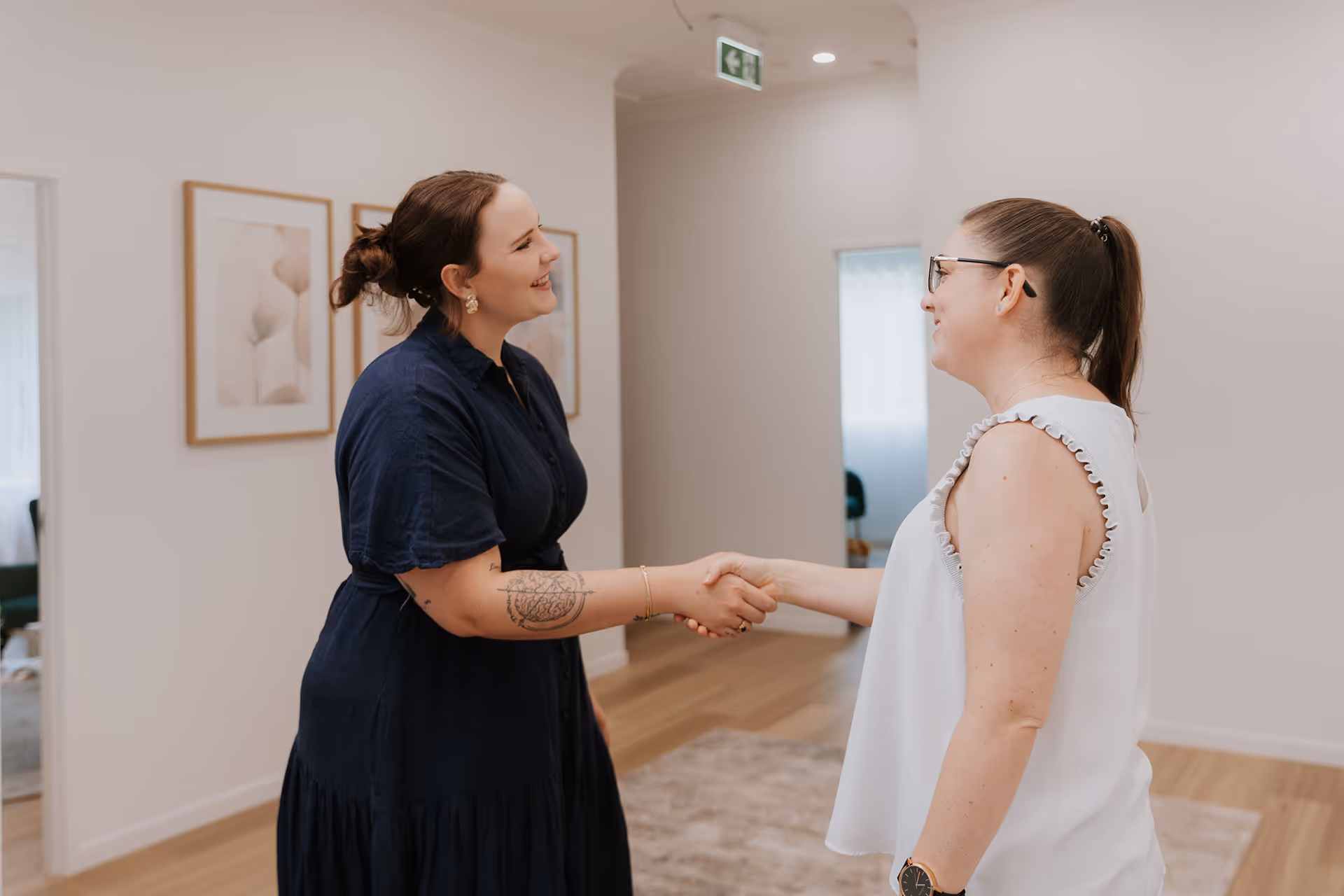 Two women smiling and shaking hands in a bright room with wooden floors and framed art on the wall.