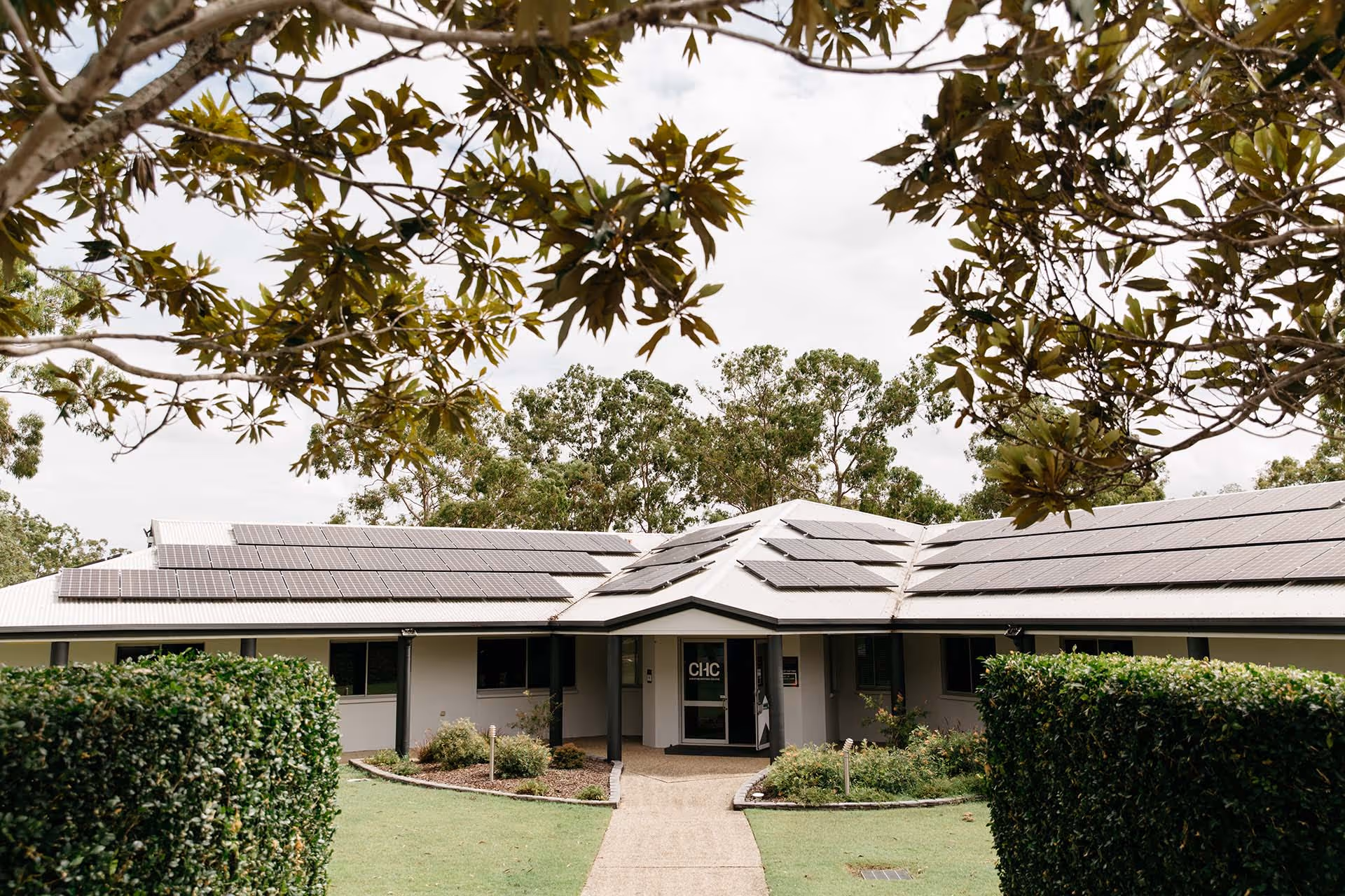 Single-story building with solar panels on the roof, surrounded by trimmed hedges and trees.
