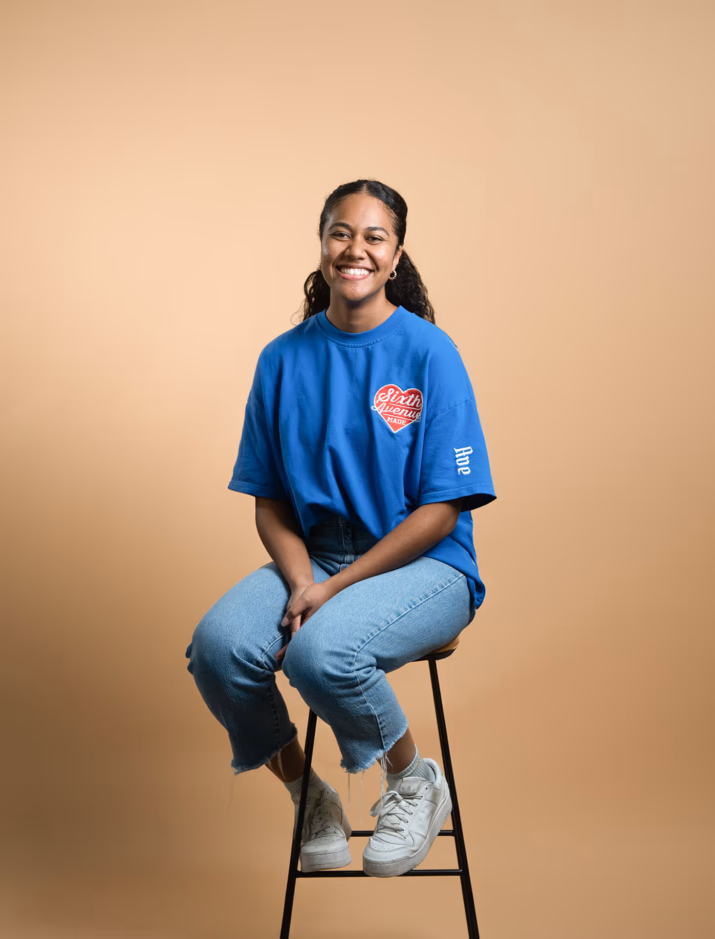Smiling woman with curly hair tied back, wearing a blue t-shirt and jeans, sitting on a tall stool against a beige background.