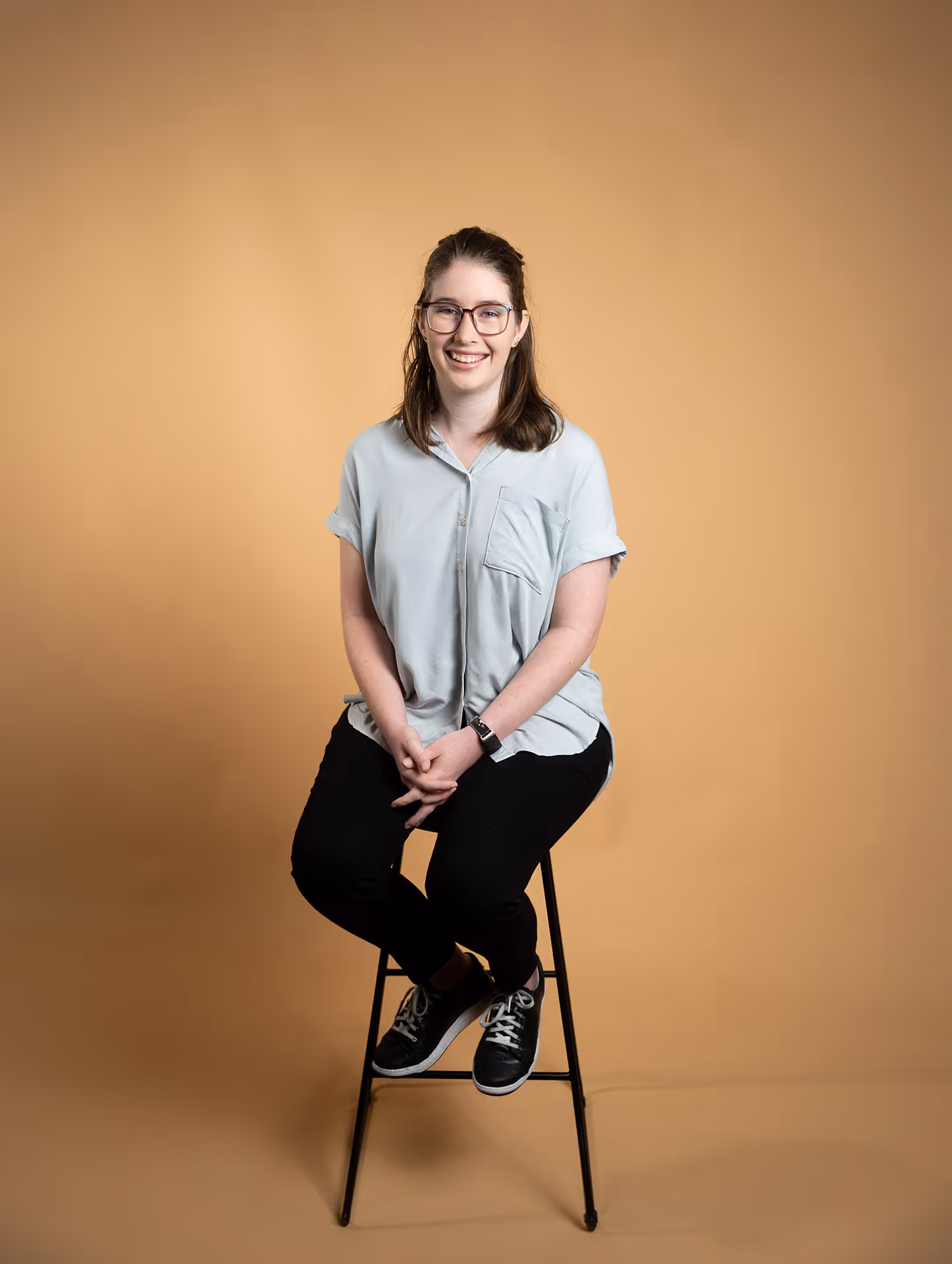Smiling woman with glasses sitting on a black stool against a beige background.