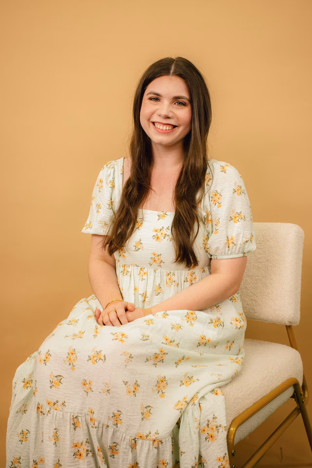 Young woman with long brown hair in a white floral dress sitting on a beige chair against a light brown background smiling.