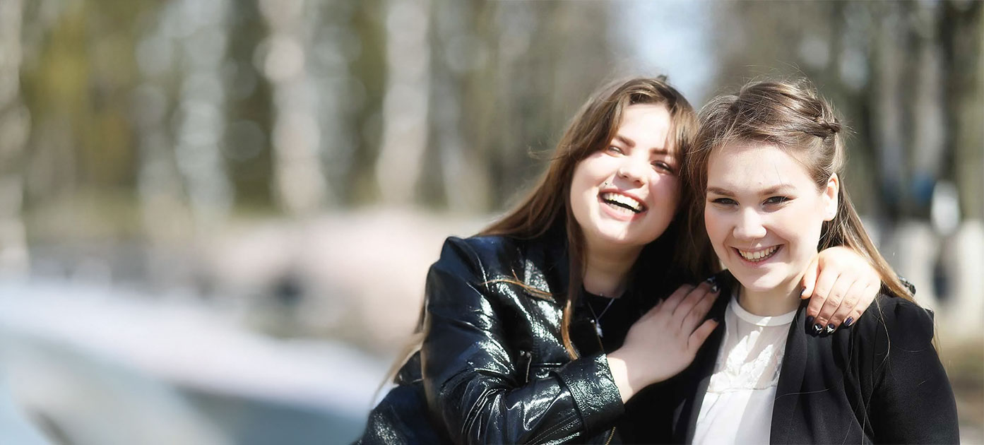 Two smiling people outside, one with an arm around the other, in sunny weather.